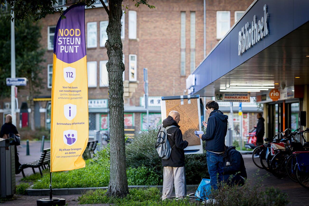 Foto van twee mannen in gesprek met elkaar voor een bibliotheek. Er hangt ook een grote vlag met de tekst noodsteunpunt